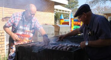 Two men cooking sausages on a grill outside.
