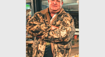 A man in a camouflage jacket standing in front of a table.