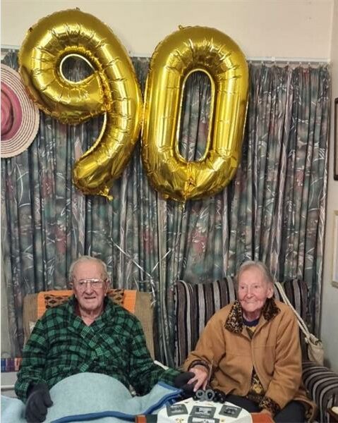 An elderly couple sitting in front of a cake with a gold balloon.
