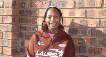 A young woman in a brown sweatshirt standing next to a brick wall.