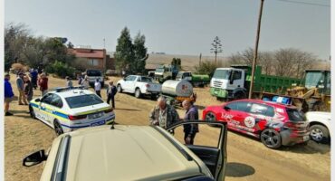Police and people standing around a car in the dirt.