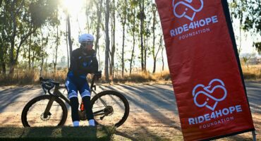 Cyclist resting next to a Ride4Hope Foundation banner during a charity cycling event in a scenic outdoor setting.