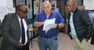 Three men standing in a room looking at papers.
