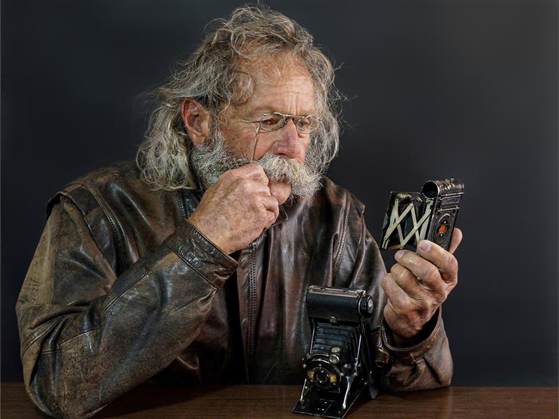 An old man with a beard and glasses holding an old camera.