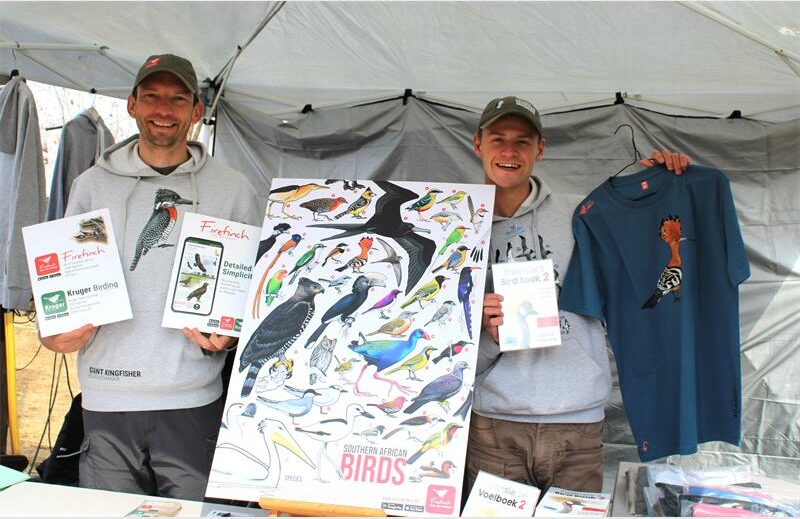 Two men standing under a tent with a bird poster.