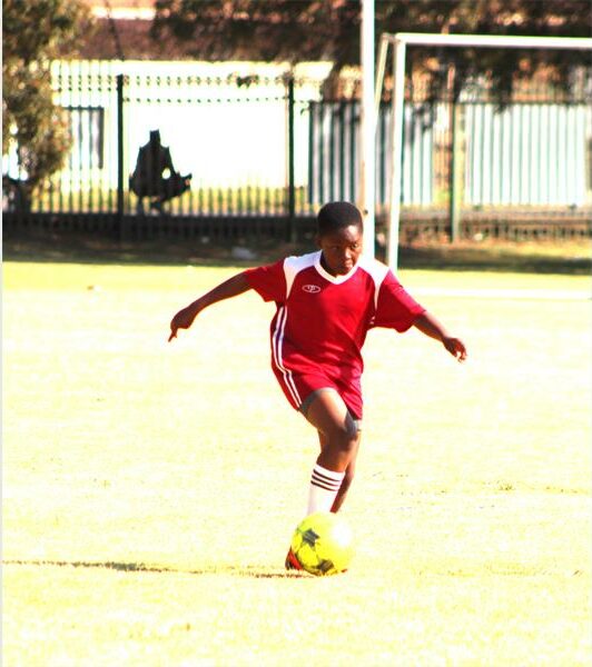 A young boy in a red uniform is kicking a soccer ball.