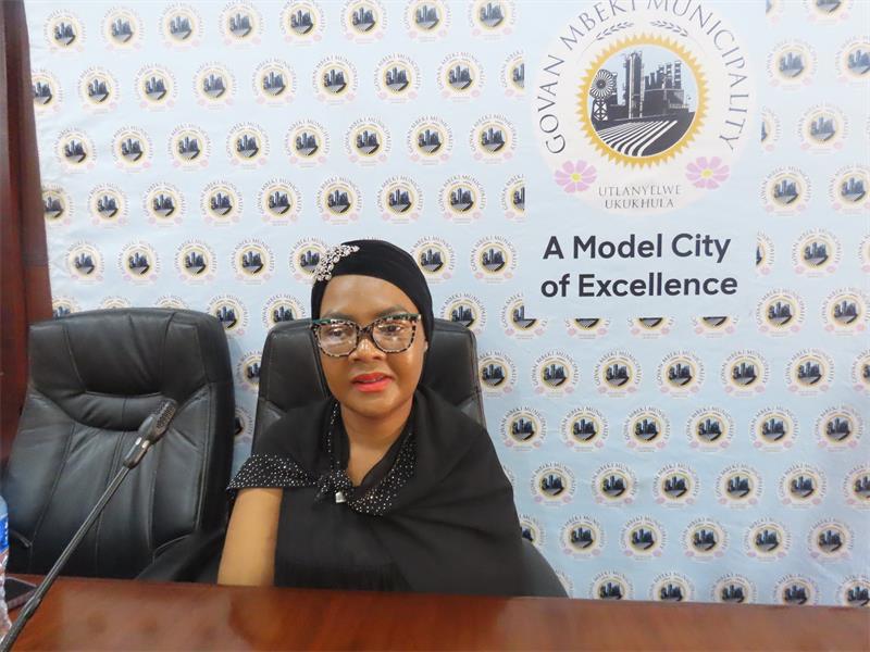 A woman in black glasses sitting at a table with a sign that says model city of excellence.