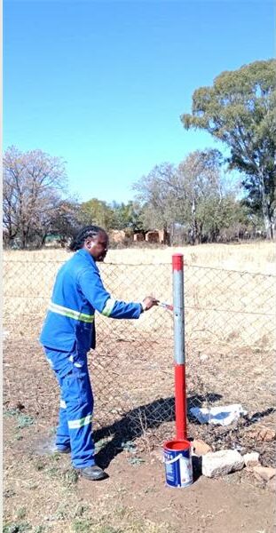 A man in blue overalls is standing next to a fence.