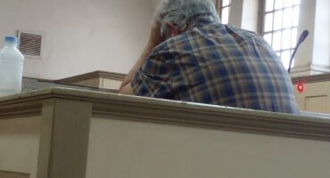 A stressed elderly man sitting at a table in a historic building, supporting his head with one hand.