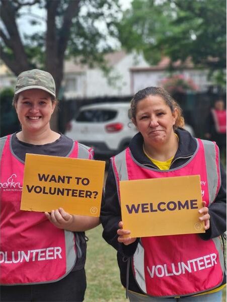 Two women in pink volunteer bibs hold up yellow signs.