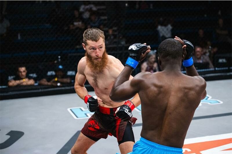 Man with red beard and boxing gloves faces opposes man in blue shorts.