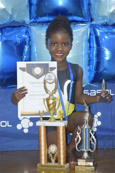 black girl in blue leotard holds up certificates and trophies