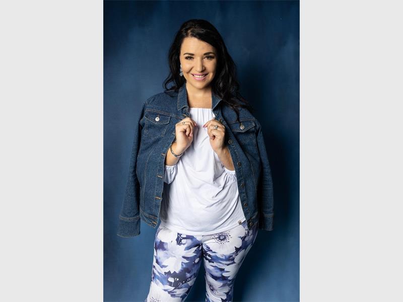Smiling woman in denim jacket and floral pants against blue background, casual fashion, Ridge Times local news feature.