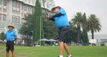 man tees off on golf course with building in back ground