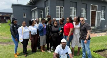 people stand in front of a grey building. one man is crouching