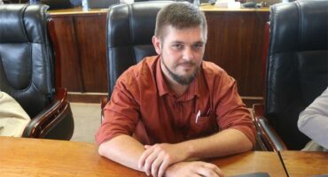 man with beard wearing rust coloured shirt sits at a desk