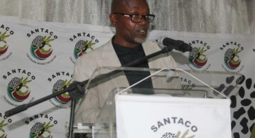 man speaks over microphone. He is standing behind a podium on which the letters SANTACO appears