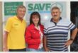 woman in red is flanked by two elderly men in front of the logo of the SAVF