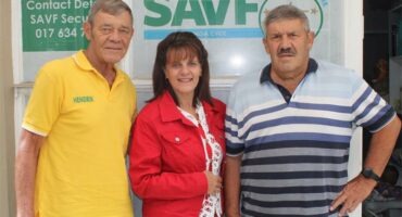 woman in red is flanked by two elderly men in front of the logo of the SAVF