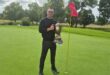 man in black standing on the golf greens with a large trophy