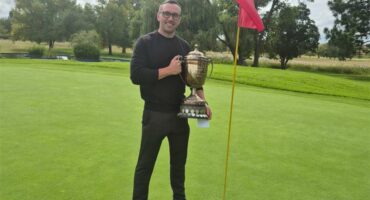 man in black standing on the golf greens with a large trophy