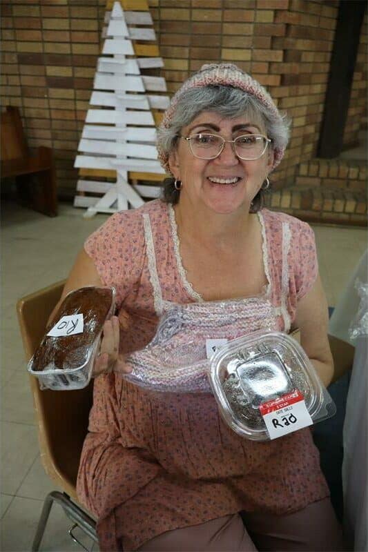 Older woman smiling, holding a cake and a dessert container, seated indoors.