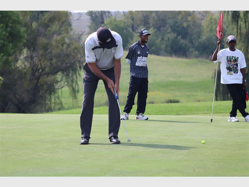 Golf players practicing putting on the green with a caddie and spectators nearby.