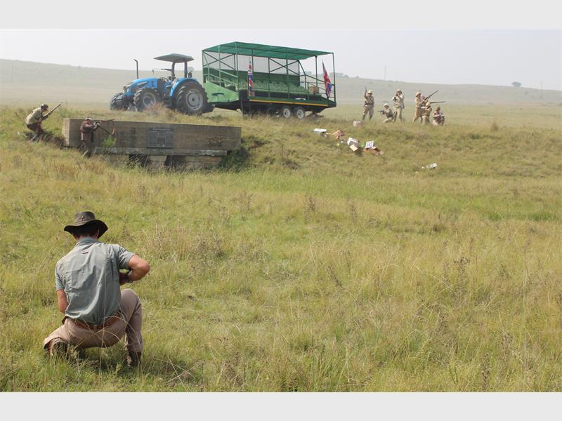 n man in boere-klere hurk in die veld. In die agtergrond is 'n trekker en mense in die klere van Britse soldate