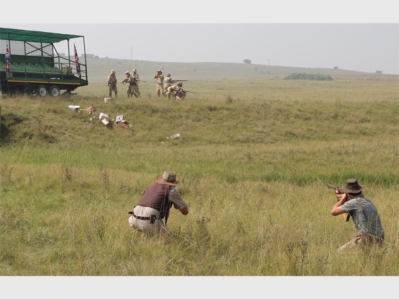 n man in boere-klere hurk in die veld. In die agtergrond is 'n trekker en mense in die klere van Britse soldate