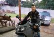 Man in a leather jacket sitting on a grey motorcycle underneath a carport
