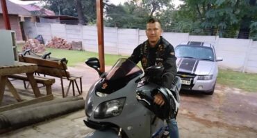 Man in a leather jacket sitting on a grey motorcycle underneath a carport
