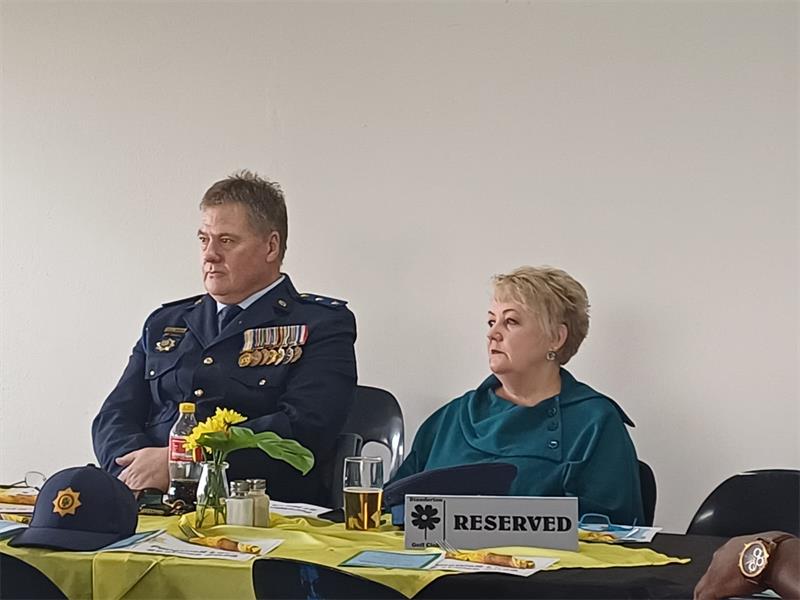 a man in a police formal uniform sitting at a table. A woman with a teal dress sits next to him,