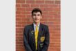 boy in black and yellow school uniform stands in front of a brick wall