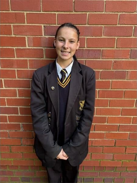girl in black and yellow school uniform stands in front of a brick wall
