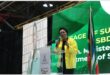 woman wearing a yellow jacket stands at a podium to speak into a microphone