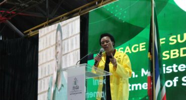 woman wearing a yellow jacket stands at a podium to speak into a microphone