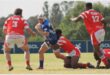 A rugby player in blue runs with the ball while the red team tries to stop him