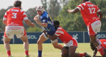 A rugby player in blue runs with the ball while the red team tries to stop him