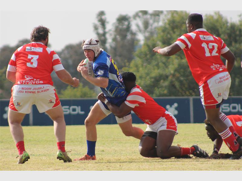 A rugby player in blue runs with the ball while the red team tries to stop him
