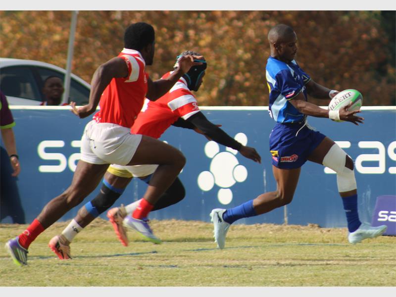 A rugby player in blue runs with the ball while the red team tries to stop him