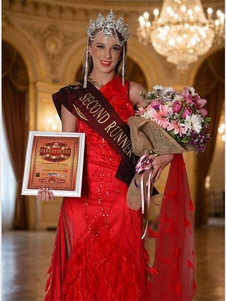young model in a red dress wears a crown. A flower bouquet is in her hand.