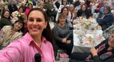a woman in pink takes a selfie with many women at a ladies tea in the background