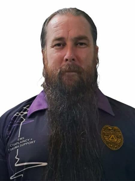 Close-up image of a man with a beard and a moustache, wearing a purple shirt, against a plain white background.