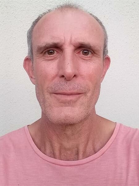 Close-up image of a elderly man with short grey hair, wearing a pink shirt, against a plain white background.