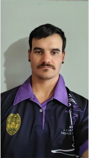 Close-up image of a young man with short brown hair and a moustache, wearing a purple shirt, against a plain white background.