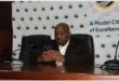 man sits at a desk with his hands together. behind him is a banner for Govan Mbeki Municipality