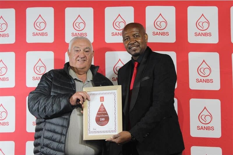 Two men hold up a certificate. The red background contains logos of the SANBS