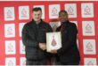 Two men hold up a certificate. The red background contains logos of the SANBS