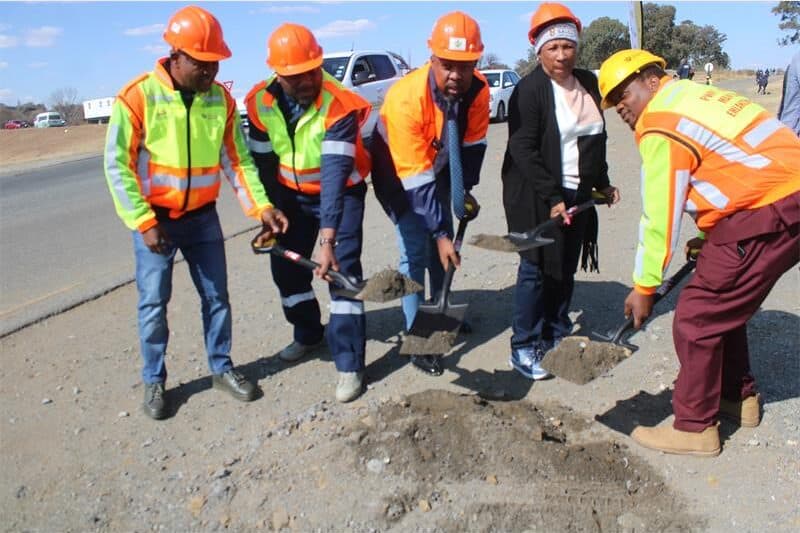five people with reflector jackets, hard hats shovel gravel