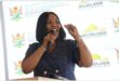 a woman speaks over a a microphone. There is a backdrop with the government logos